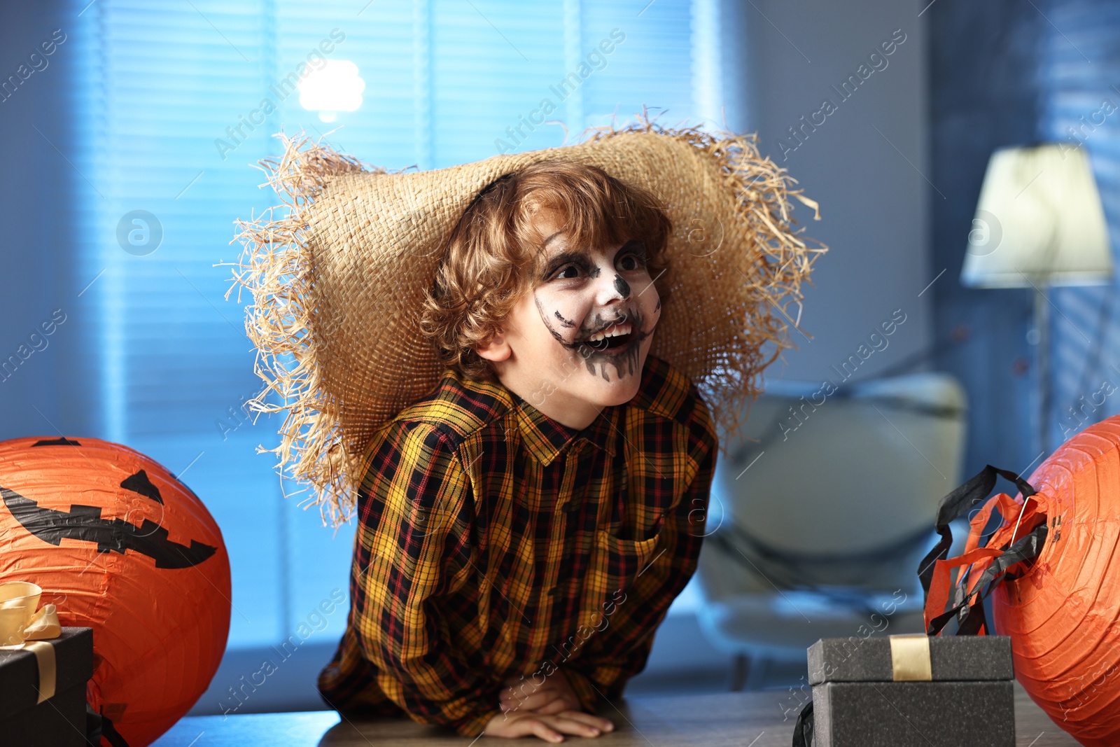 Photo of Cute boy dressed like scarecrow with festive decor and gift boxes indoors at night. Halloween celebration