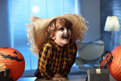 Cute boy dressed like scarecrow with festive decor and gift boxes indoors at night. Halloween celebration