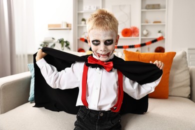 Funny boy dressed like vampire for Halloween celebration on sofa in room