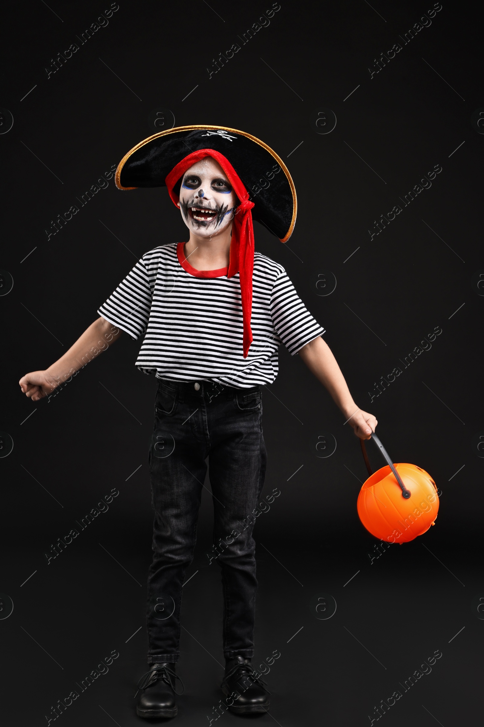 Photo of Funny boy with pumpkin bucket dressed like pirate on black background. Halloween costume