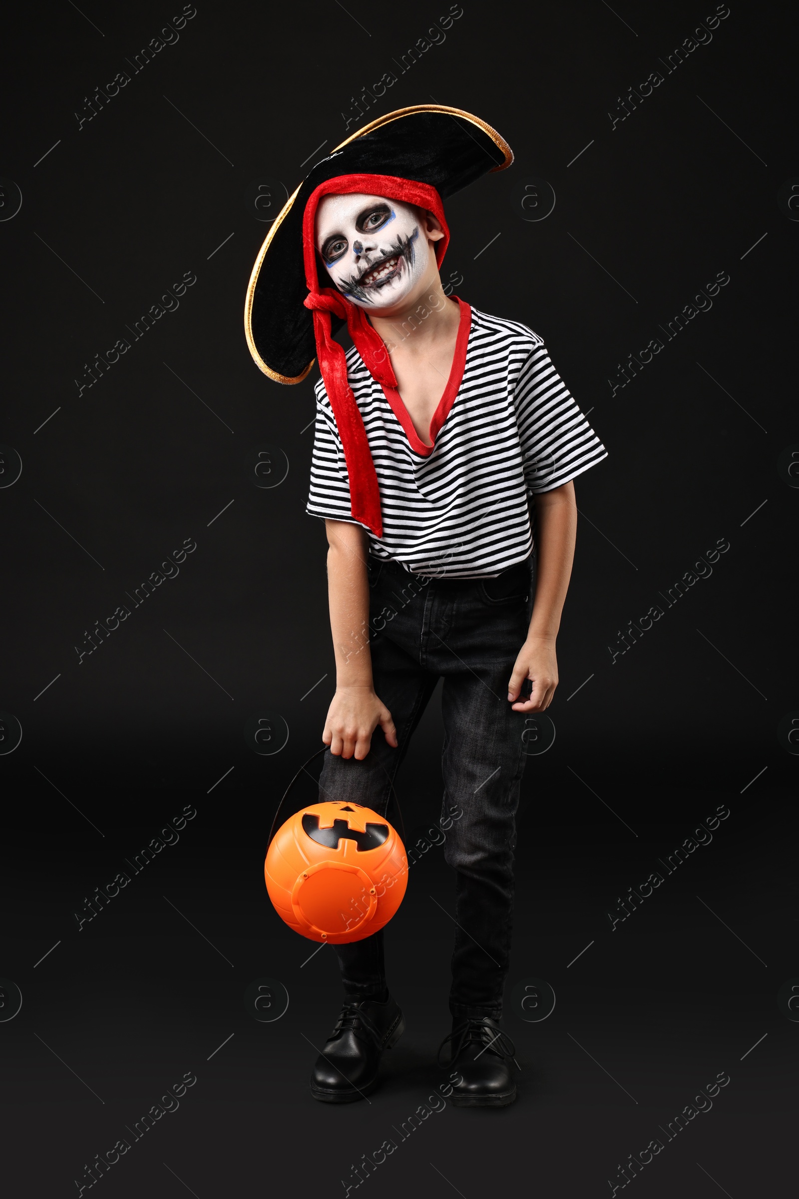 Photo of Funny boy with pumpkin bucket dressed like pirate on black background. Halloween costume