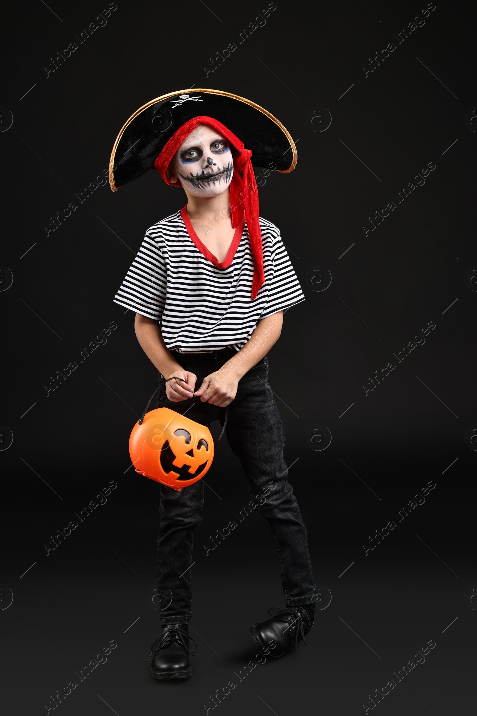 Photo of Funny boy with pumpkin bucket dressed like pirate on black background. Halloween costume