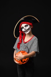 Photo of Funny boy with pumpkin bucket dressed like pirate on black background. Halloween costume