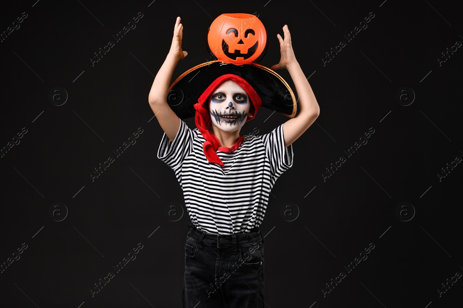 Photo of Funny boy with pumpkin bucket dressed like pirate on black background. Halloween costume