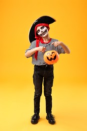 Photo of Funny boy with pumpkin bucket dressed like pirate on yellow background. Halloween costume