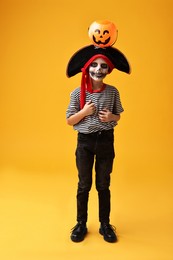 Photo of Funny boy with pumpkin bucket dressed like pirate on yellow background. Halloween costume