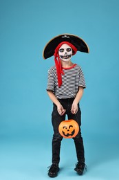 Photo of Funny boy with pumpkin bucket dressed like pirate on light blue background. Halloween costume