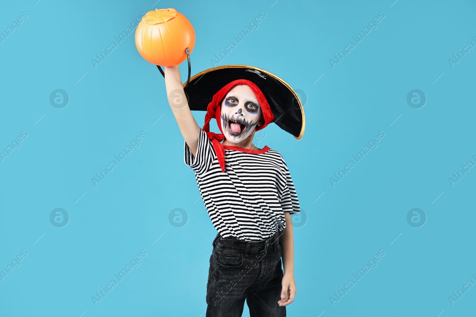 Photo of Funny boy with pumpkin bucket dressed like pirate on light blue background. Halloween costume