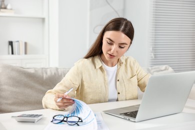 Photo of Budget planning. Young woman with papers using laptop at white table indoors