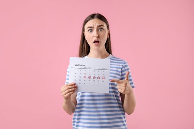 Emotional woman holding calendar with marked menstrual cycle days on pink background