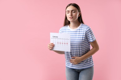 Woman holding calendar with marked menstrual cycle days and suffering from abdominal pain on pink background, space for text