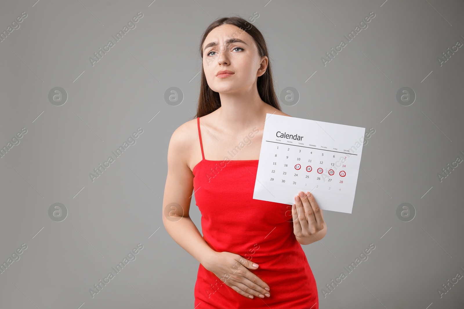 Photo of Woman holding calendar with marked menstrual cycle days and suffering from abdominal pain on grey background