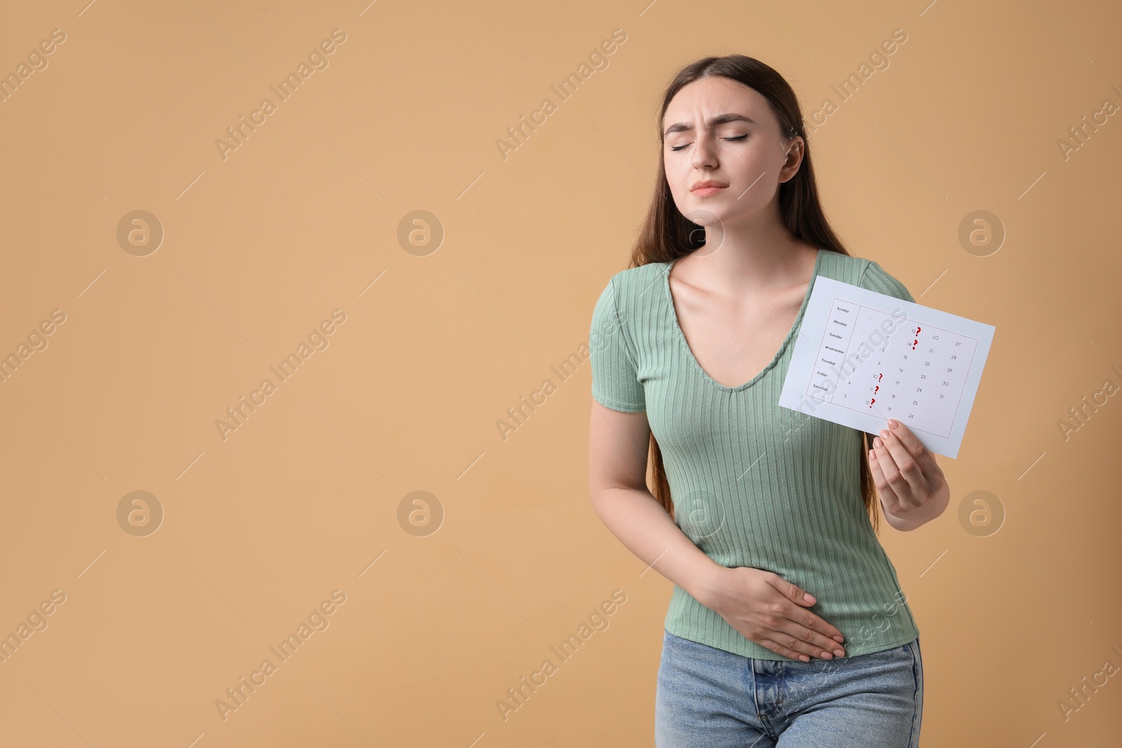 Photo of Woman holding calendar with marked menstrual cycle days and suffering from abdominal pain on beige background, space for text