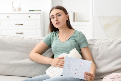 Photo of Woman holding calendar with marked menstrual cycle days and using hot water bottle on sofa at home