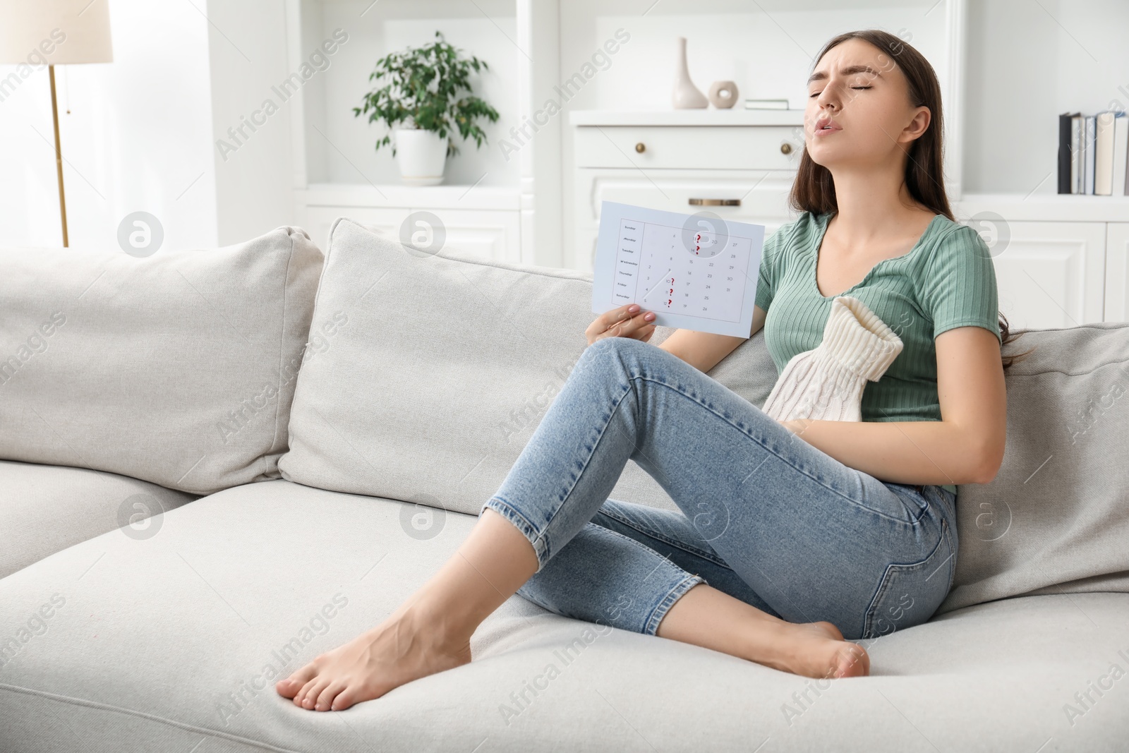 Photo of Woman holding calendar with marked menstrual cycle days and using hot water bottle on sofa at home