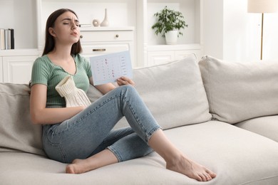 Photo of Woman holding calendar with marked menstrual cycle days and using hot water bottle on sofa at home