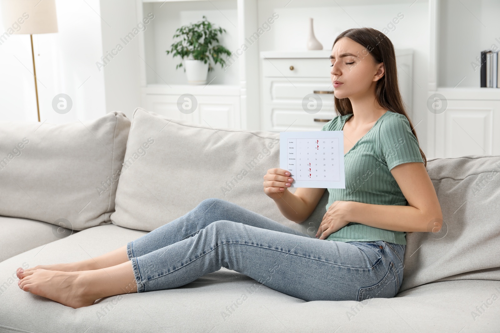 Photo of Woman holding calendar with marked menstrual cycle days and suffering from abdominal pain on sofa at home