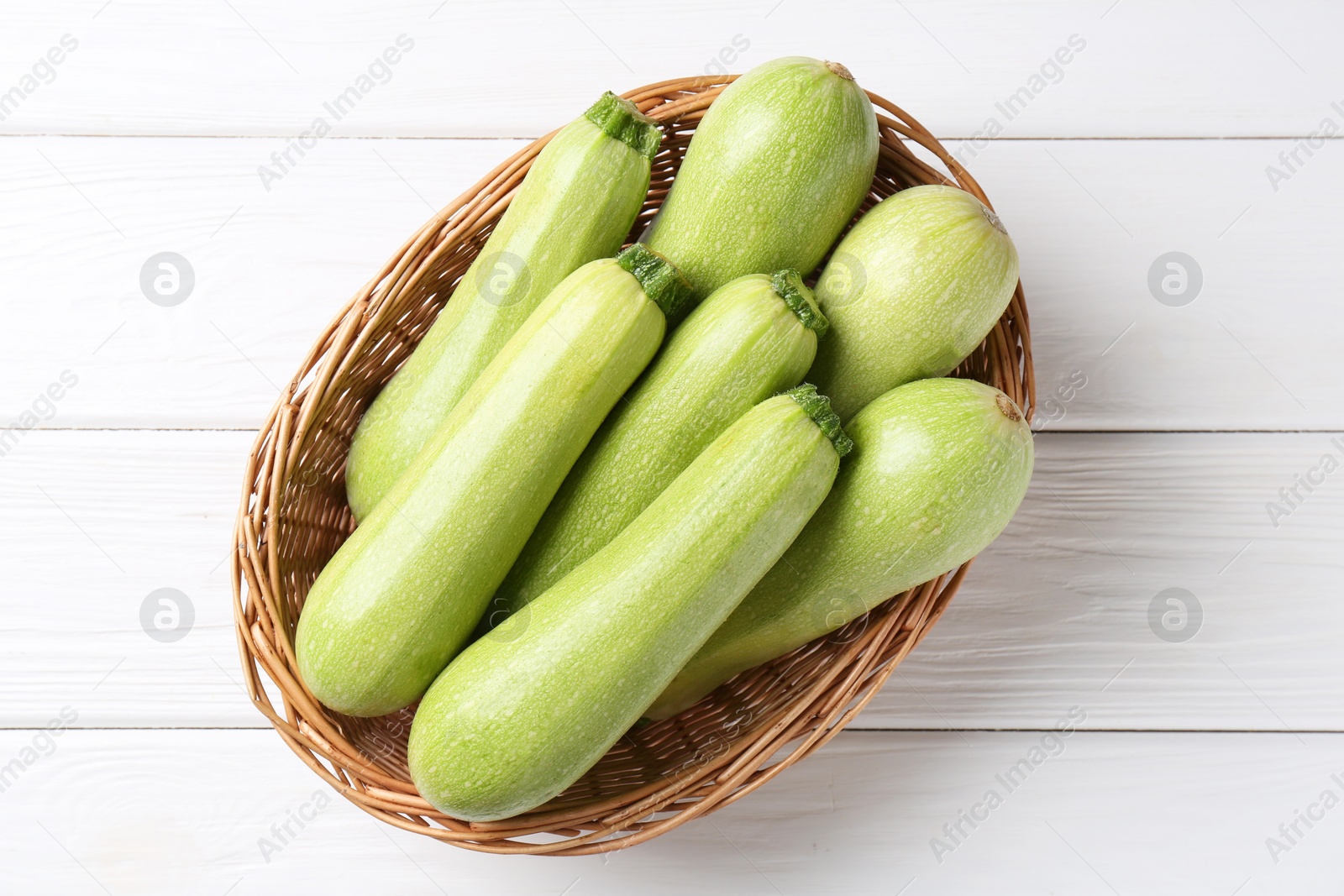 Photo of Fresh zucchinis in wicker basket on white wooden table, top view