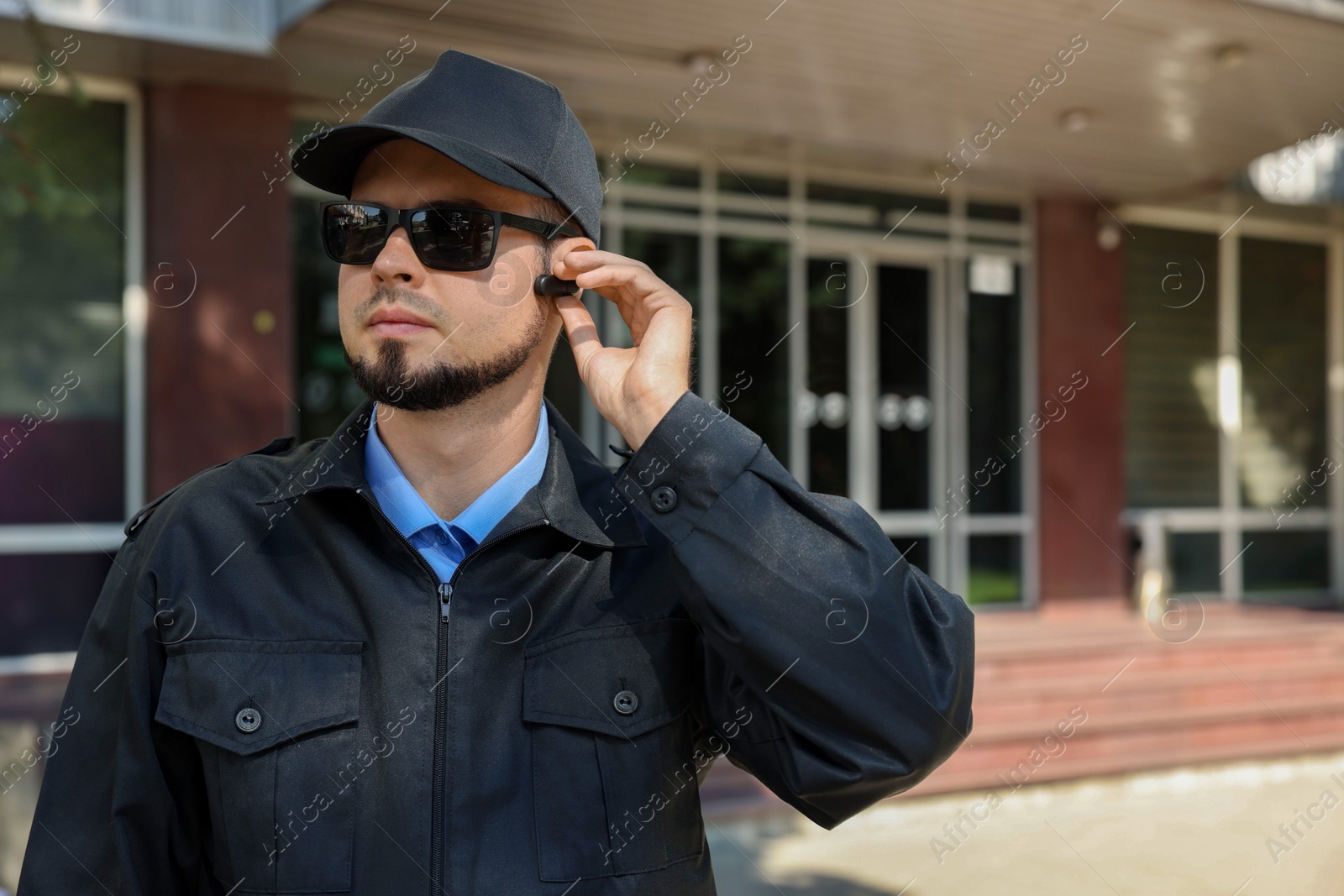 Photo of Security guard in uniform and sunglasses using earpiece outdoors