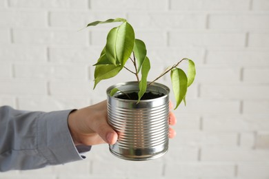 Photo of Recycling concept. Woman holding metal can with growing plant against white brick wall, closeup