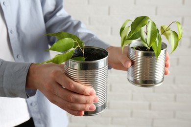 Photo of Recycling concept. Woman holding metal cans with growing plants against white brick wall, closeup