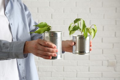 Photo of Recycling concept. Woman holding metal cans with growing plants against white brick wall, closeup