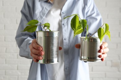 Photo of Recycling concept. Woman holding metal cans with growing plants against white brick wall, closeup