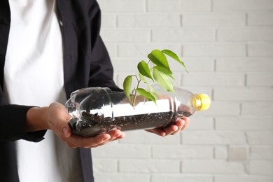 Photo of Recycling concept. Woman holding plastic bottle with growing plant against white brick wall, closeup