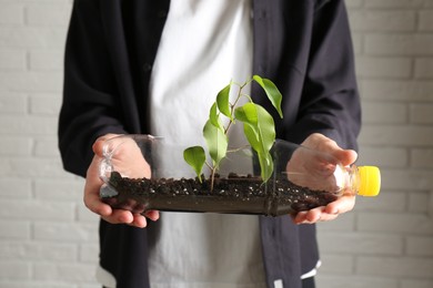 Photo of Recycling concept. Woman holding plastic bottle with growing plant against white brick wall, closeup