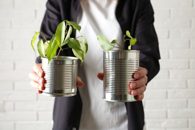 Photo of Recycling concept. Woman holding metal cans with growing plants against white brick wall, closeup