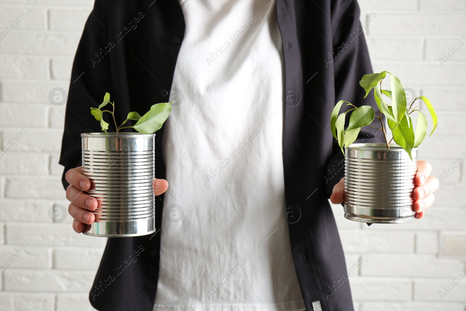 Photo of Recycling concept. Woman holding metal cans with growing plants against white brick wall, closeup