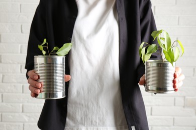 Photo of Recycling concept. Woman holding metal cans with growing plants against white brick wall, closeup