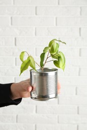 Photo of Recycling concept. Woman holding metal can with growing plant against white brick wall, closeup