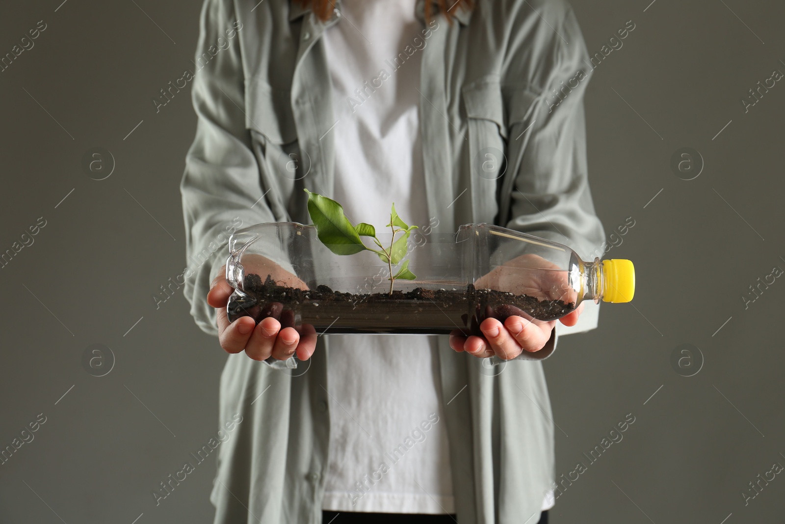 Photo of Recycling concept. Woman holding plastic bottle with growing plant against grey background, closeup