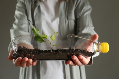 Photo of Recycling concept. Woman holding plastic bottle with growing plant against grey background, closeup