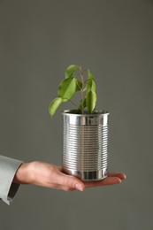 Photo of Recycling concept. Woman holding metal can with growing plant against grey background, closeup