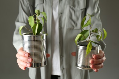 Photo of Recycling concept. Woman holding metal cans with growing plants against grey background, closeup