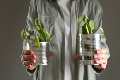 Photo of Recycling concept. Woman holding metal cans with growing plants against grey background, closeup