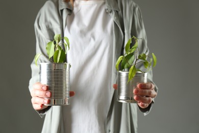 Photo of Recycling concept. Woman holding metal cans with growing plants against grey background, closeup