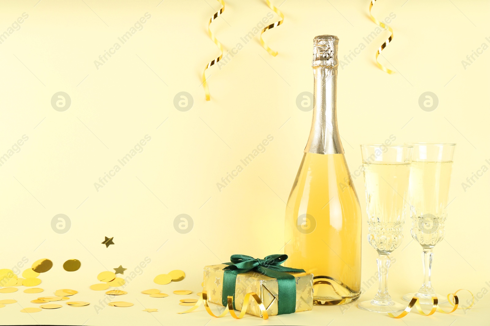 Photo of Shiny confetti, streamers, sparkling wine and gift box on beige background. Space for text