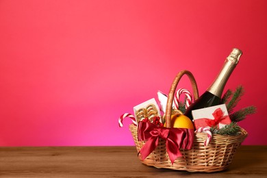 Photo of Wicker gift basket with wine on wooden table against pink background, space for text