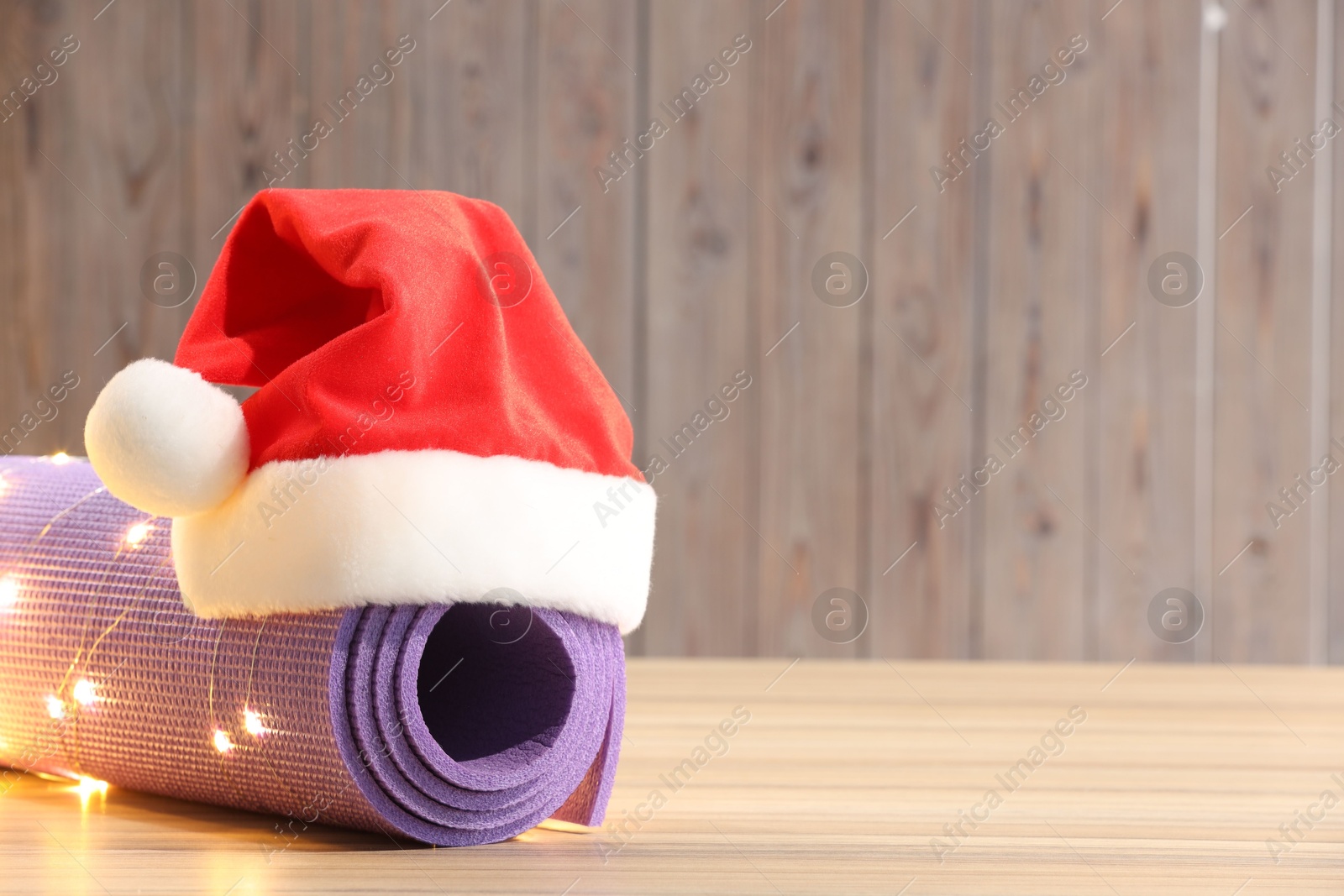 Photo of Yoga mat with Christmas lights and Santa hat on wooden surface. Space for text