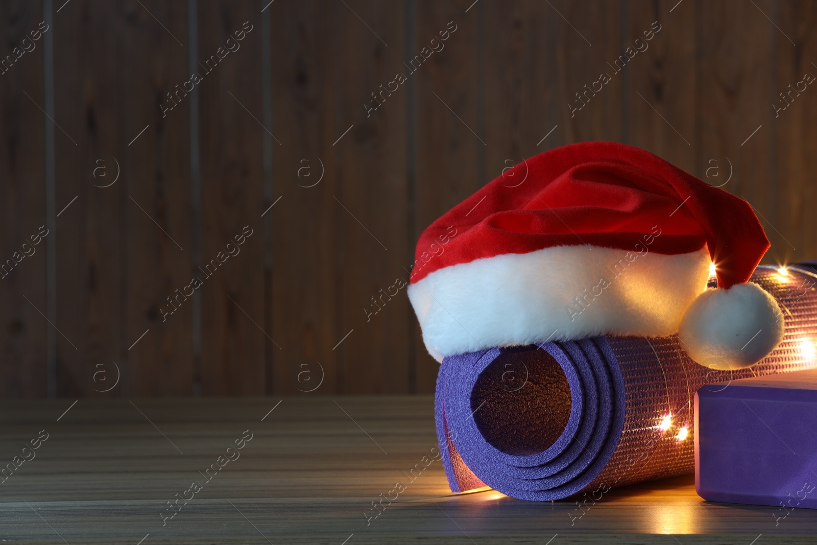 Photo of Yoga mat with Christmas lights and Santa hat on wooden surface. Space for text