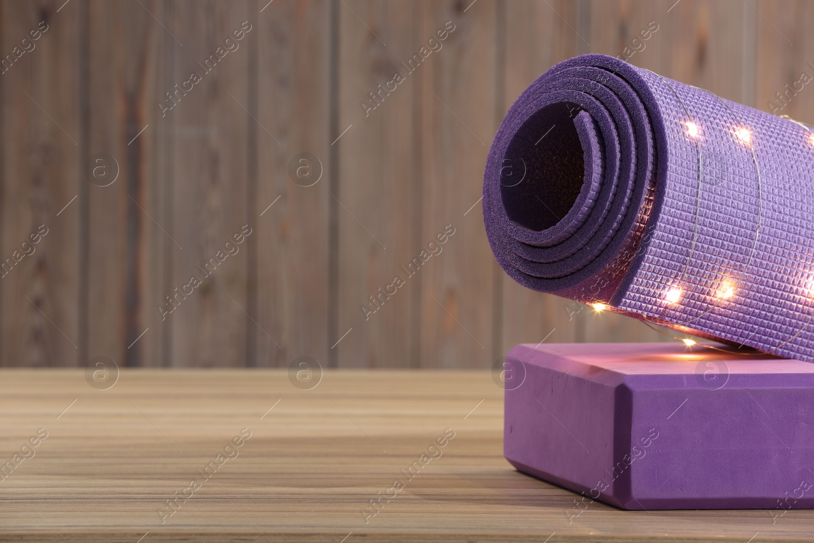 Photo of Yoga mat and block with Christmas lights on wooden surface. Space for text