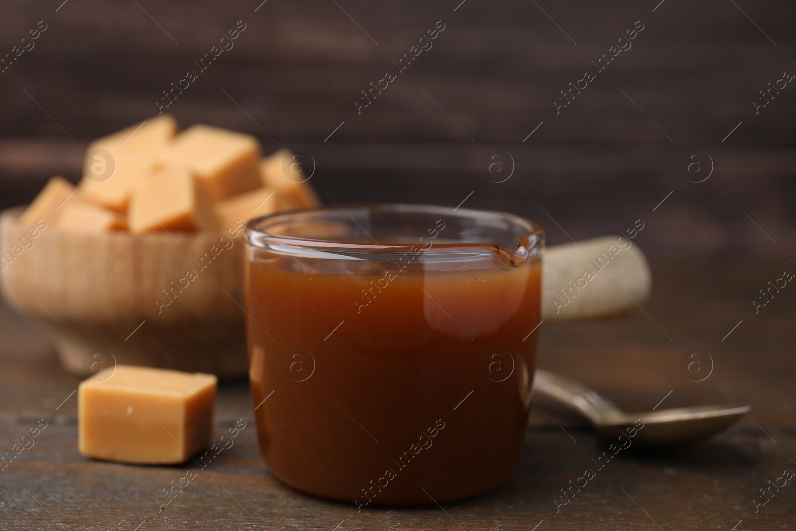 Photo of Tasty caramel sauce in glass measuring cup and sweet candies on wooden table, closeup