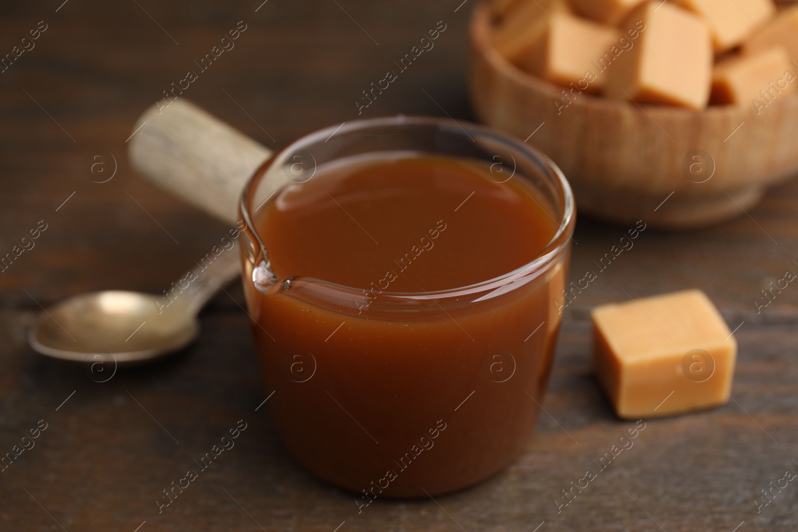 Photo of Tasty caramel sauce in glass measuring cup and sweet candies on wooden table, closeup