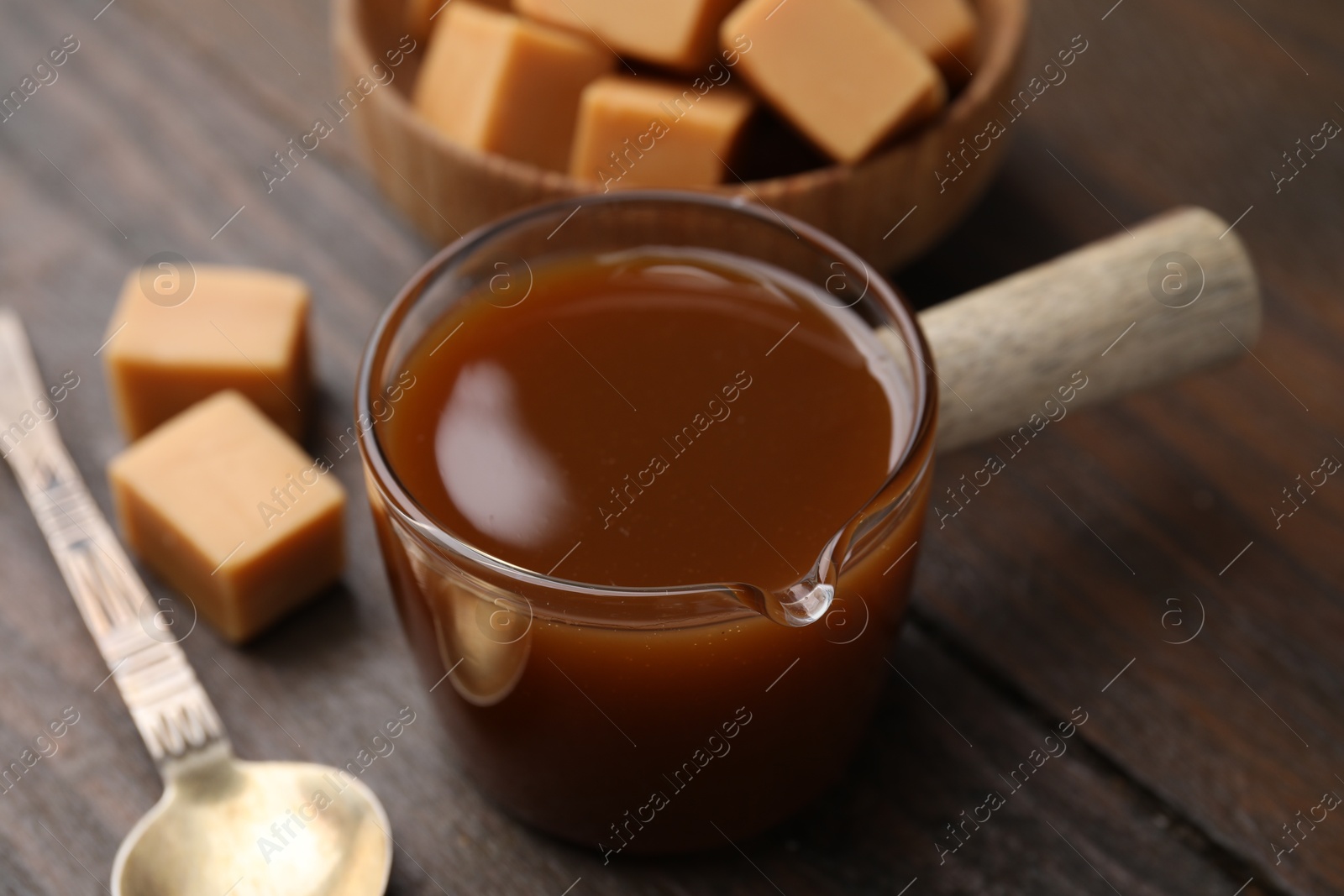 Photo of Tasty caramel sauce in glass measuring cup, sweet candies and spoon on wooden table, closeup