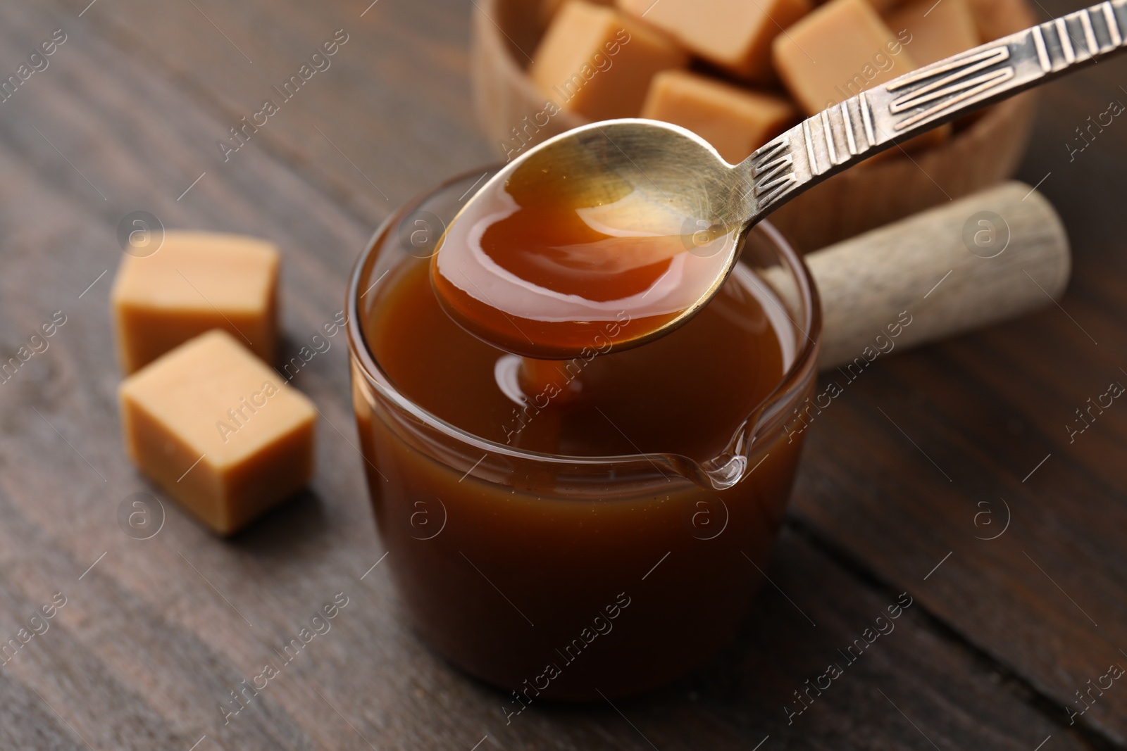 Photo of Taking tasty caramel sauce with spoon from glass measuring cup at wooden table, closeup