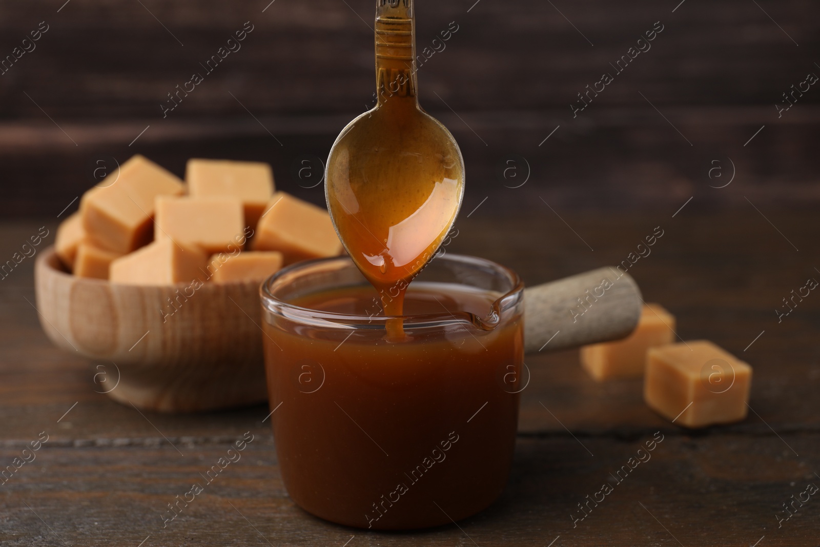 Photo of Taking tasty caramel sauce with spoon from glass measuring cup at wooden table, closeup