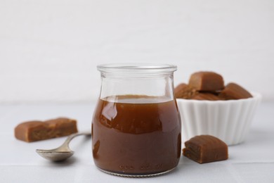 Photo of Tasty caramel sauce in glass jar, sweet candies and spoon on light table, closeup